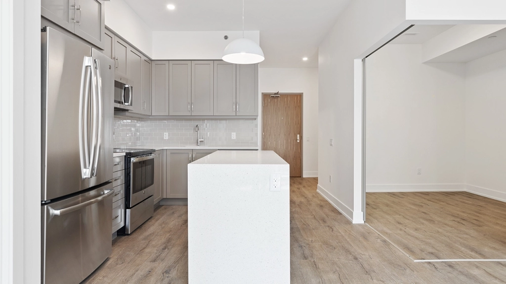 Interior image of a condo showing the front door, a kitchen island, a bedroom and open concept kitchen.