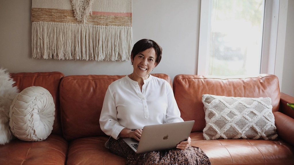 An image of a senior, mature widow lady sitting on her brown leather couch with an Apple laptop on her lap planning on downsizing for retirement.